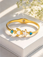 A gold-plated bangle bracelet with a fish and starfish motif, accented by blue stones, displayed on a white box with soft floral shadows.