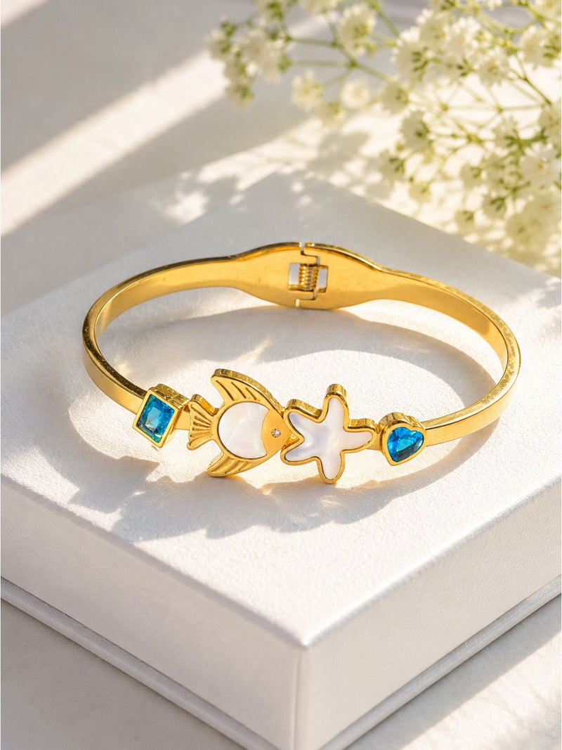 A gold-plated bangle bracelet with a fish and starfish motif, accented by blue stones, displayed on a white box with soft floral shadows.