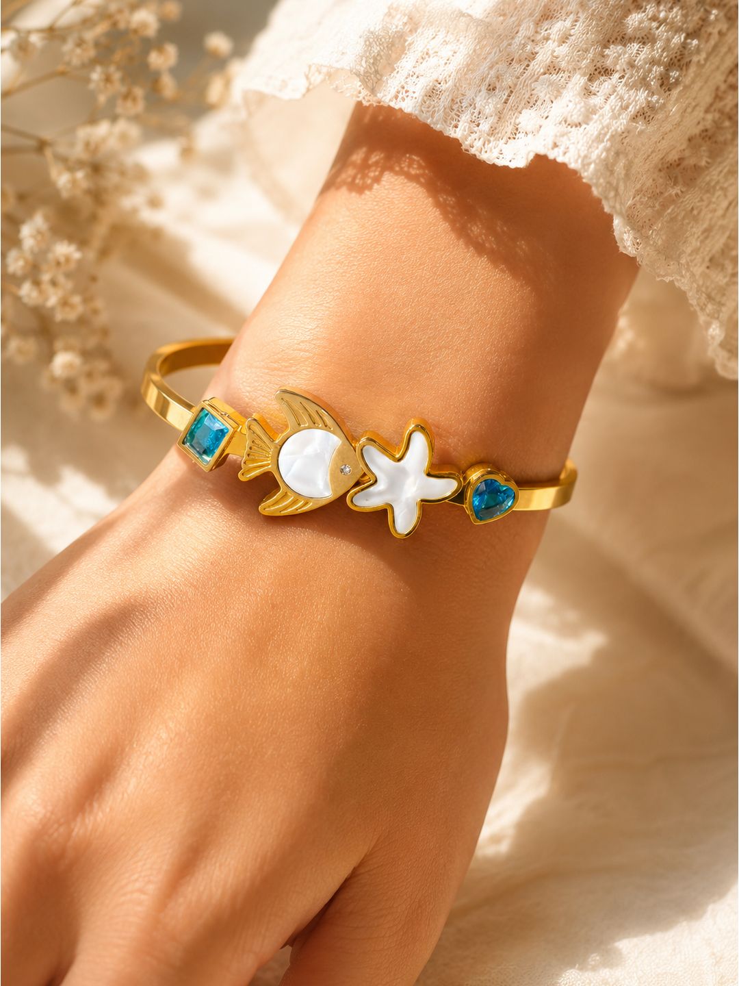 A gold-toned cuff bracelet with sea-themed charms including a crescent moon, starfish, and fish, accented with blue and white stones, shown worn on a wrist.