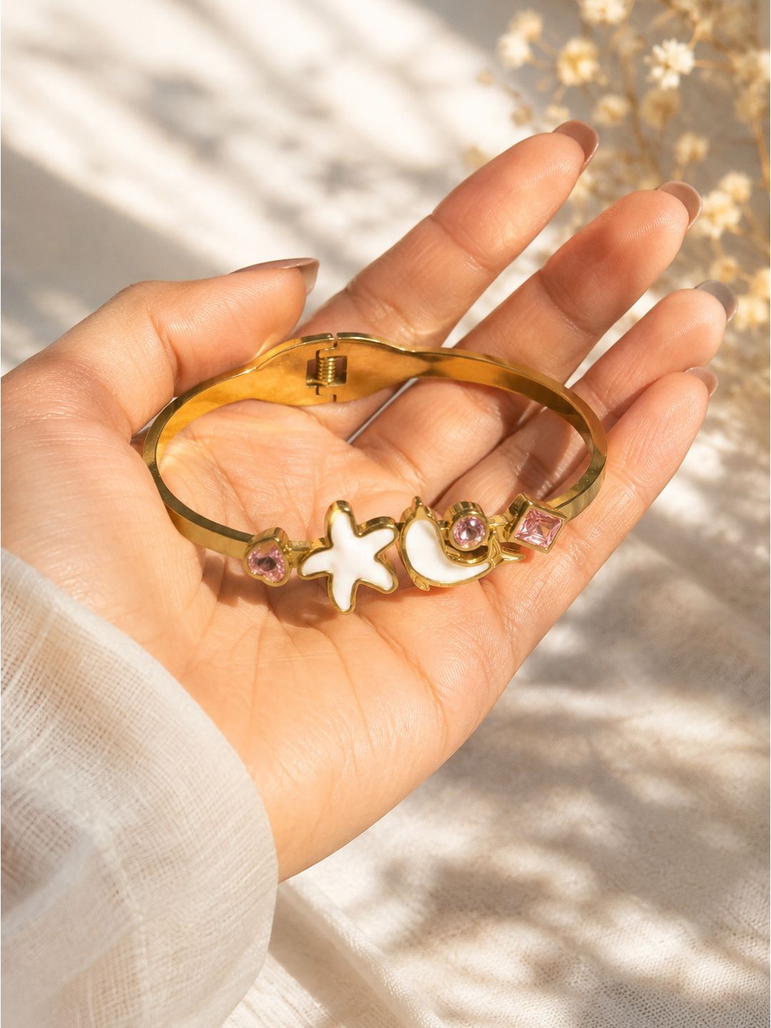 A gold bangle bracelet with a starfish and crescent moon charm, displayed on a person's hand against a softly lit, sun-dappled background.