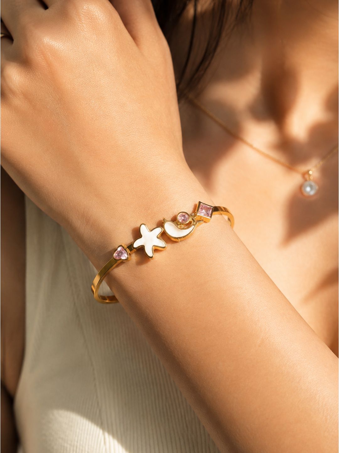 A gold bangle bracelet with a starfish, crescent moon, and pink gemstone charms, shown worn on a woman's wrist in warm sunlight.