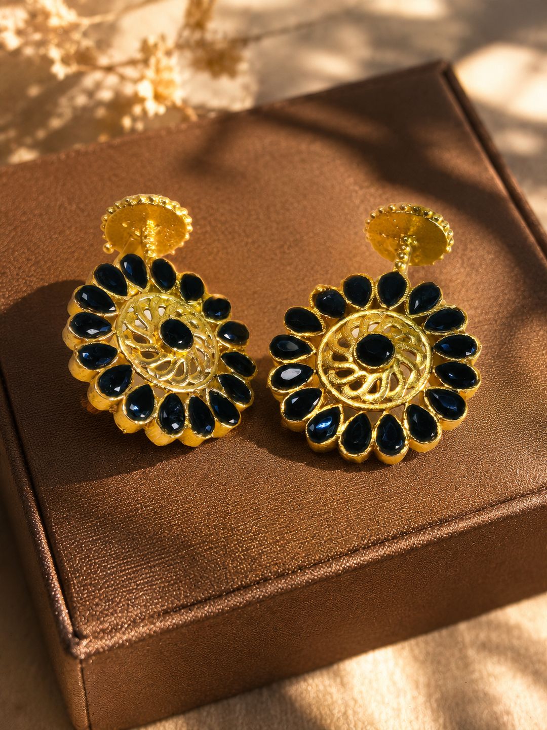 A pair of gold-toned statement earrings with black teardrop-shaped stones arranged in a sunburst pattern, displayed on a brown textured box.