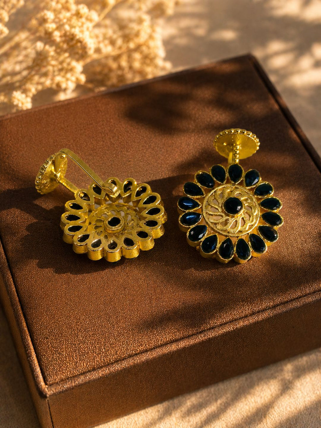 A pair of gold-toned floral earrings with dark blue teardrop stones and a central black stone, displayed on a brown jewelry box with warm sunlight and shadows.