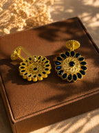 A pair of gold-toned floral earrings with dark blue teardrop stones and a central black stone, displayed on a brown jewelry box with warm sunlight and shadows.