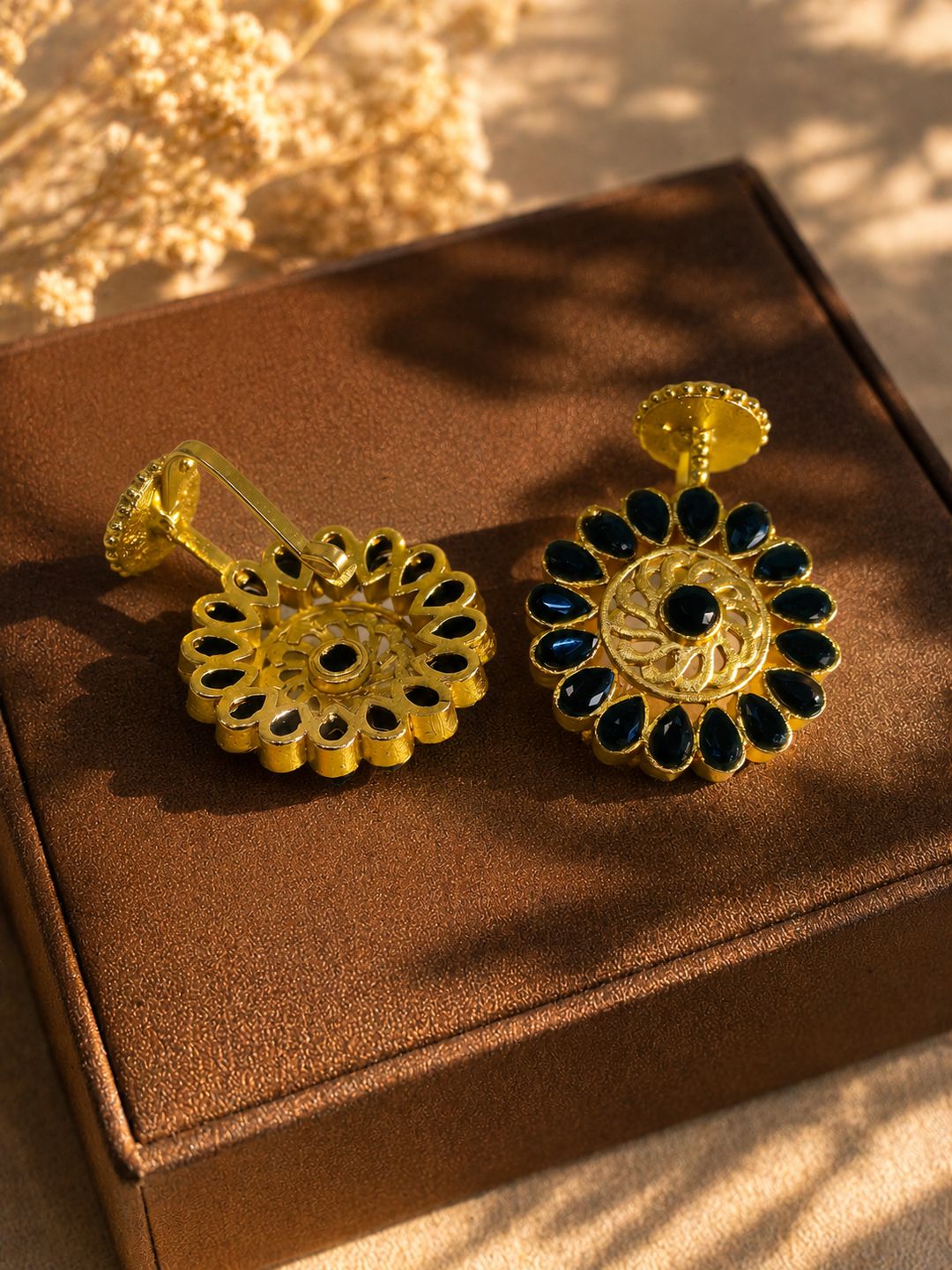 A pair of gold-toned floral earrings with dark blue teardrop stones and a central black stone, displayed on a brown jewelry box with warm sunlight and shadows.