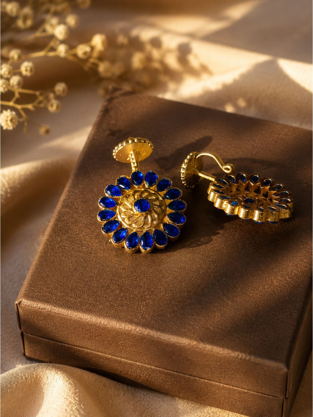 A pair of gold-toned dangle earrings with deep blue gemstone floral detailing, displayed on a brown satin box with soft beige fabric and dried flowers in the background.