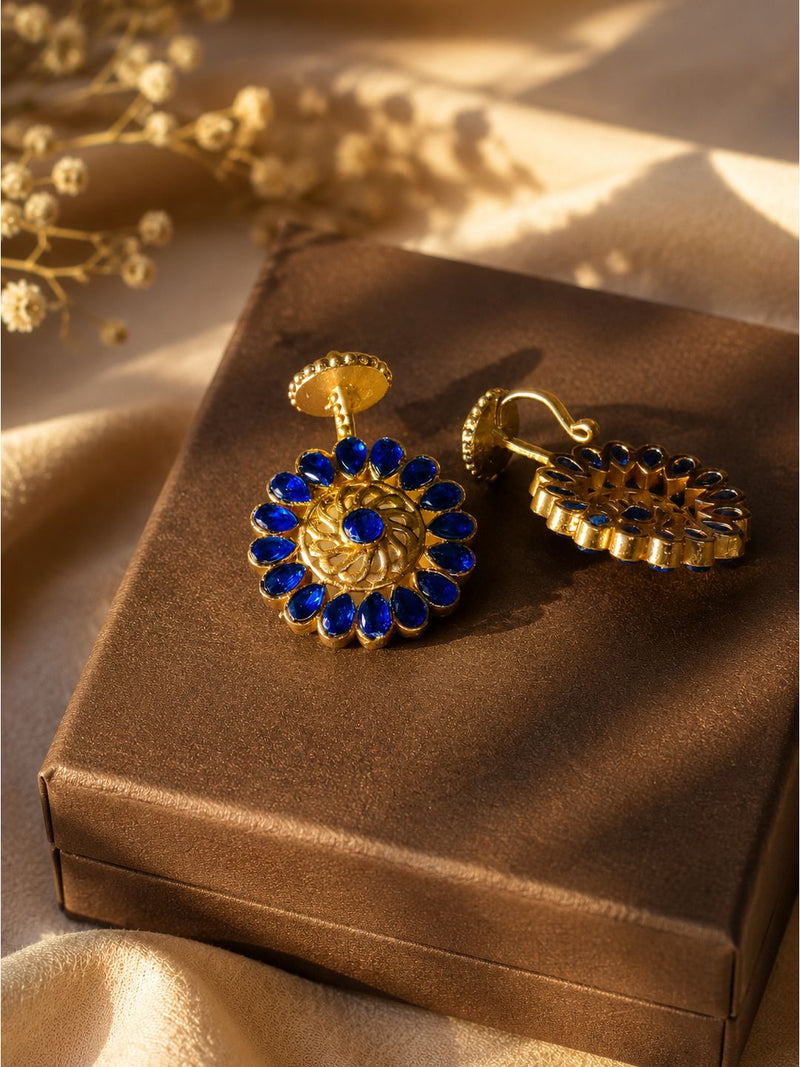 A pair of gold-toned dangle earrings with deep blue gemstone floral detailing, displayed on a brown satin box with soft beige fabric and dried flowers in the background.