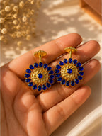A pair of gold-toned statement earrings with deep blue teardrop stones arranged in a sunburst pattern, held in a person's hand against a warm beige background with soft shadows.