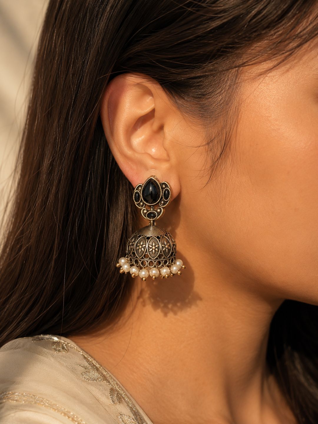 A close-up of a woman's ear wearing an ornate traditional Indian jhumka earring. The earring features a black teardrop-shaped stone in a gold-toned setting, with a dangling filigree bell adorned with small white pearl beads at the bottom.