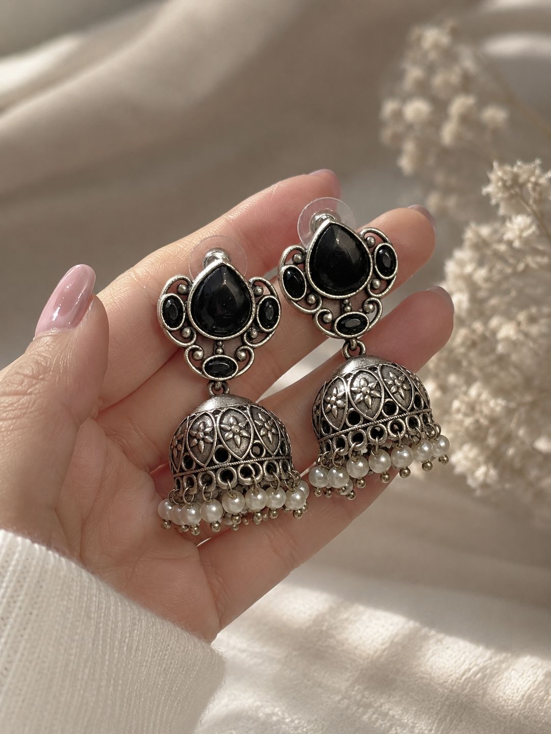 A pair of ornate silver-toned jhumka earrings with black teardrop stones and small white pearl bead drops, held in a hand against a soft beige fabric background.