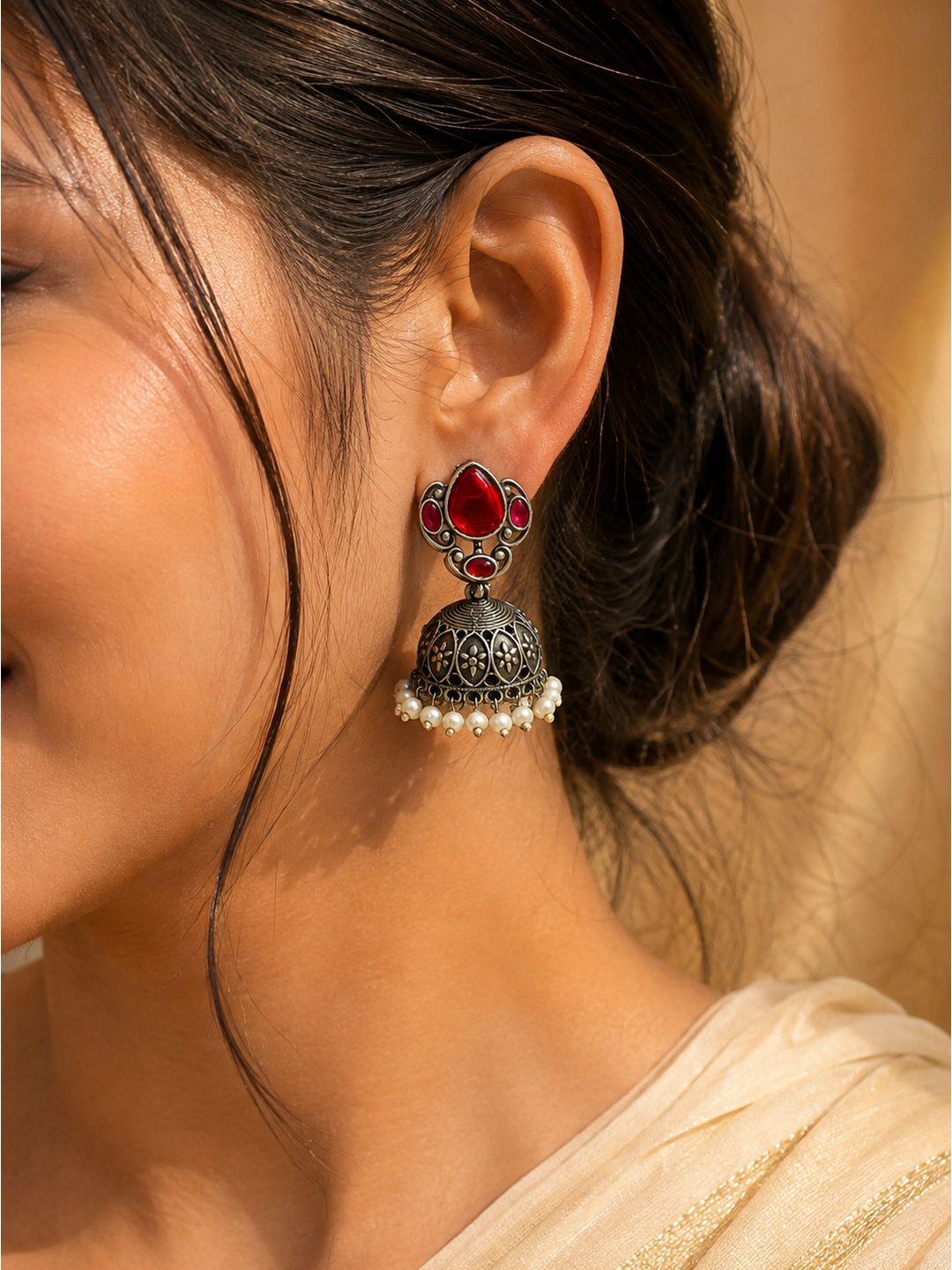 A close-up of a woman's ear wearing a traditional Indian jhumka earring. The earring features a silver-toned filigree bell with a row of small white pearls at the bottom, a silver-toned post, and a red teardrop-shaped stone and smaller red stones at the top.