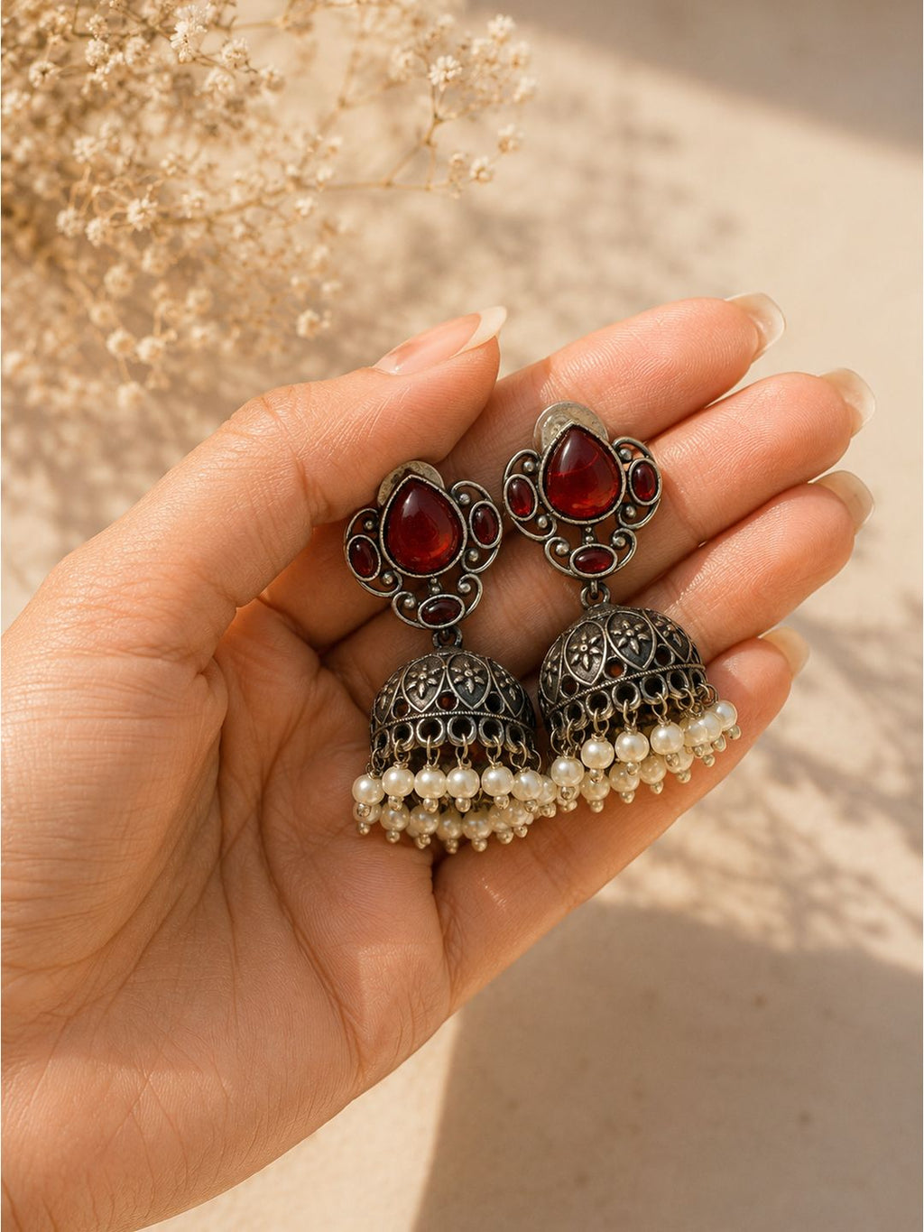 A pair of silver-toned filigree jhumka earrings with red teardrop stones and white pearl bead drops, held in a hand against a warm beige background with dried flowers.