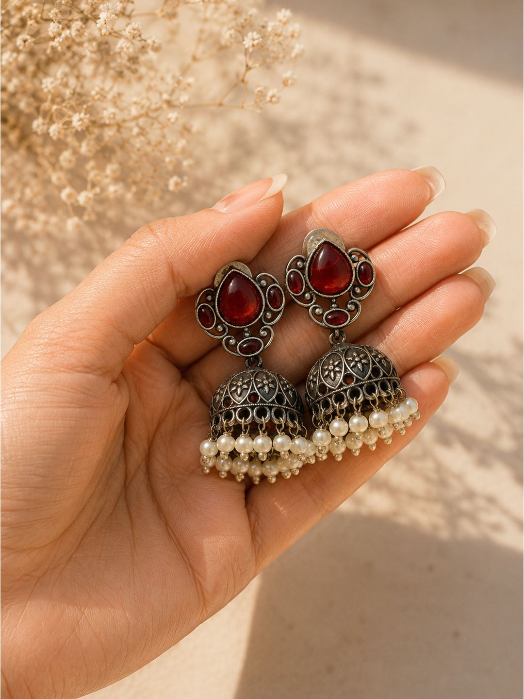 A pair of silver-toned filigree jhumka earrings with red teardrop stones and white pearl bead drops, held in a hand against a warm beige background with dried flowers.