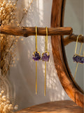 A pair of gold drop earrings with rough purple amethyst gemstones, hanging from a rustic wooden branch in front of a wooden mirror.