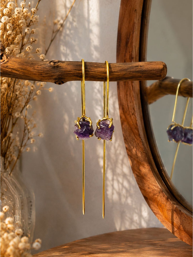 A pair of gold drop earrings with rough purple amethyst gemstones, hanging from a rustic wooden branch in front of a wooden mirror.