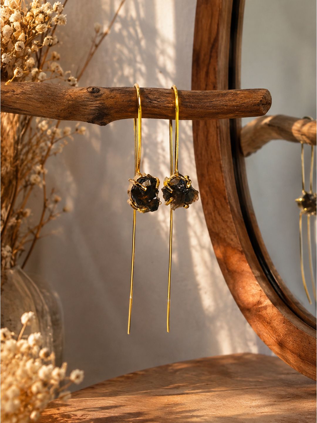 Gold-tone dangle earrings with dark textured floral clusters, hanging from a rustic wooden branch in a warm, styled setting with dried flowers and a wooden mirror frame.